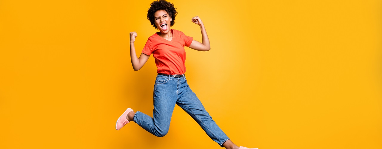 Full length photo of crazy ecstatic girl raise her fists scream shout yeah celebrate victory lottery discounts jump wear red t-shirt trendy denim jeans sneakers isolated over yellow color background